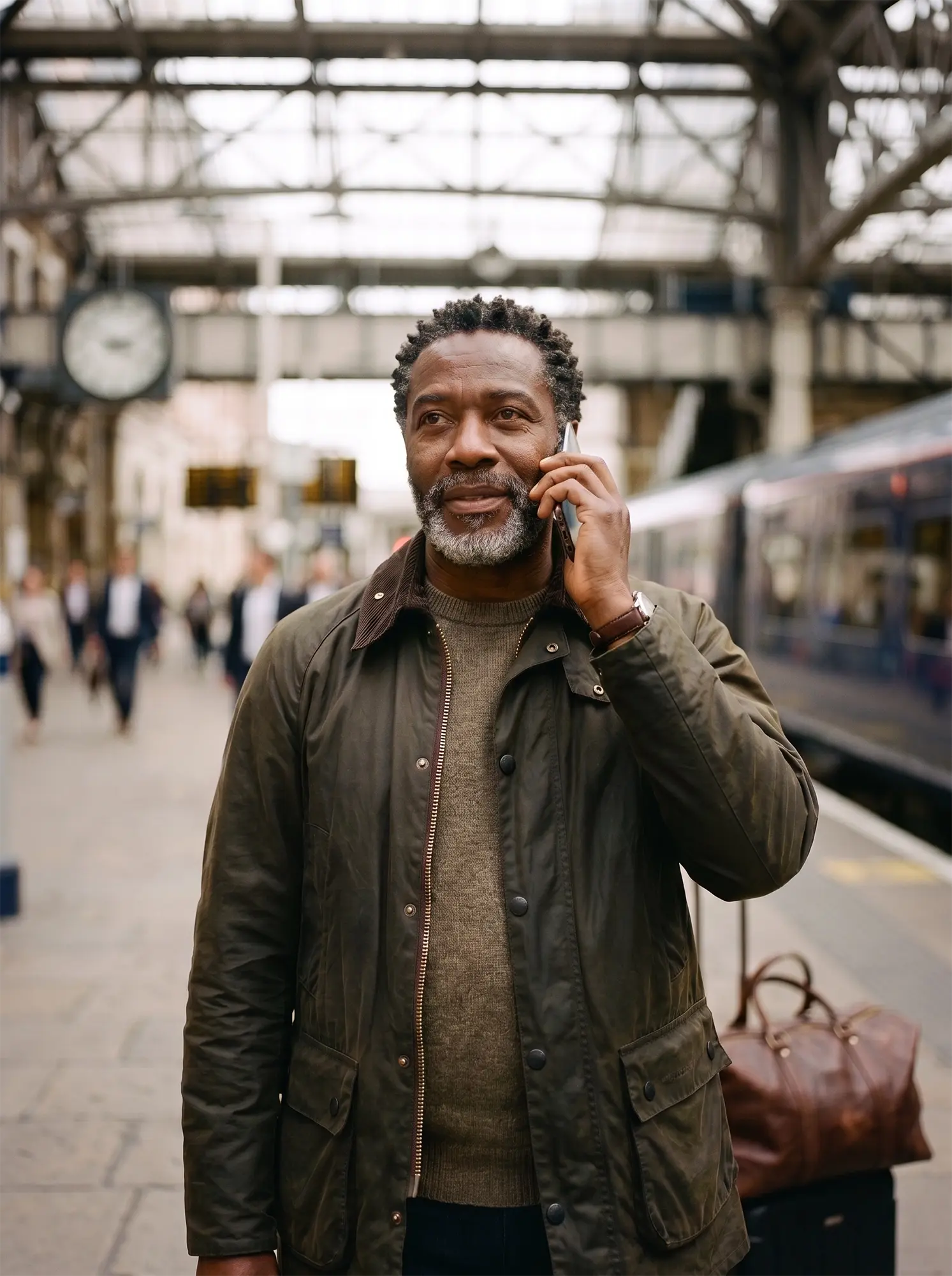 A smiling man with a beard stands at a train station while talking on his phone.
