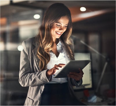A woman in a blazer smiles while using a tablet in a modern office setting.