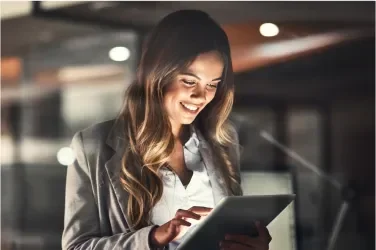 A woman in a blazer smiles while using a tablet in a modern office setting.