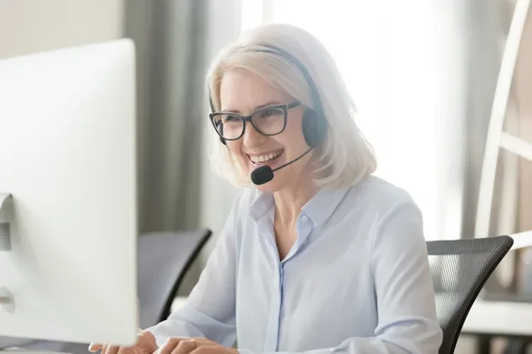 A woman with glasses smiling at her computer. She is wearing a headset