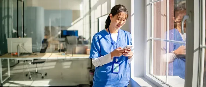 A healthcare worker in blue scrubs smiles while using a smartphone near a window.