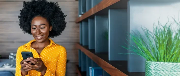 Woman in a yellow blouse smiles while using a smartphone near a bookshelf and plants.