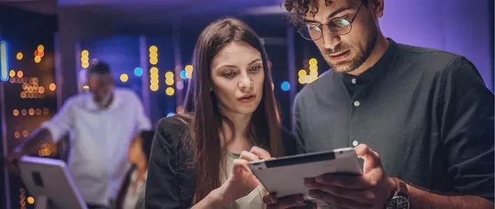 A woman and a man closely examine a tablet screen in a modern, dimly lit room.