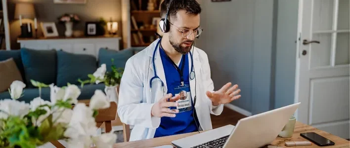 Doctor in a white coat with headphones, talking to a laptop in a cozy home office.
