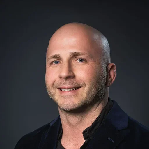 Smiling man with a bald head, wearing a dark blazer, against a dark background.