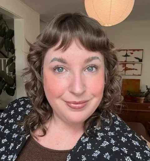 Woman with curly hair smiles warmly, wearing a patterned black jacket, indoors with soft lighting.