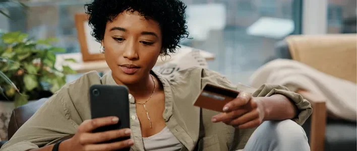 A person with curly hair holds a phone and a credit card, looking focused.