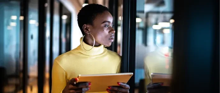 Woman in a yellow turtleneck holds a tablet, looking thoughtfully in a modern office setting.