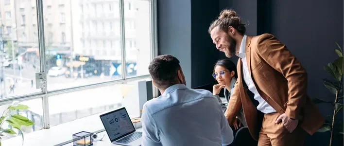 Three professionals collaborate in a bright office, with a laptop and city view.
