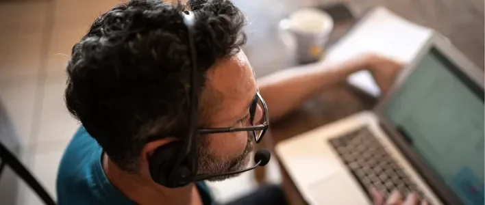 Man with curly hair and glasses wearing a headset, working on a laptop at a table.
