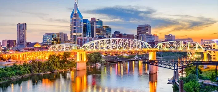 Nashville skyline at sunset, with illuminated buildings and a bridge over the river.