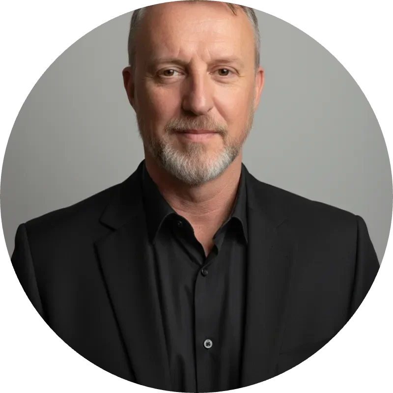 A man with a beard wearing a black suit and shirt, smiling against a gray background.