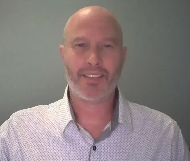 Smiling man with a bald head and beard, wearing a patterned shirt against a gray background.