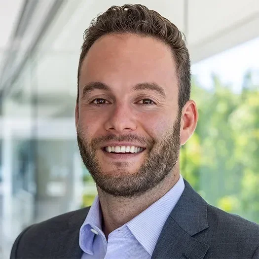 Smiling man in a suit with short hair, standing in a modern office setting.