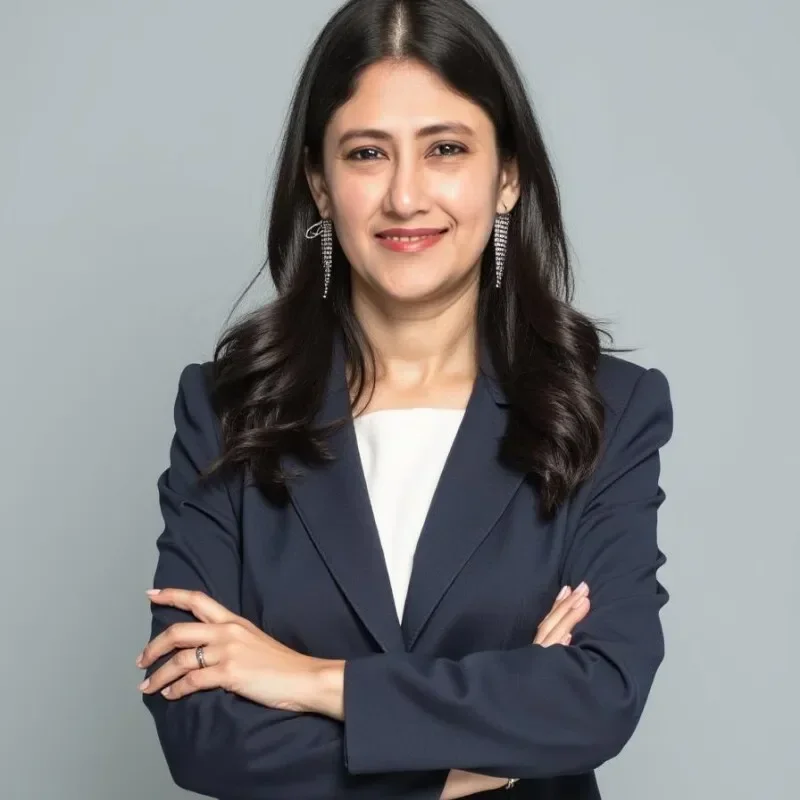 A woman in a dark blazer smiles confidently with arms crossed against a gray background.