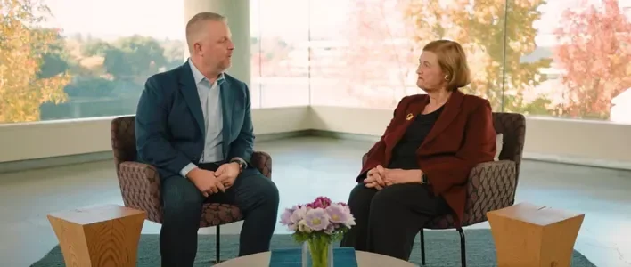 Two people sit in modern chairs, engaged in conversation, with a colorful backdrop of autumn trees.