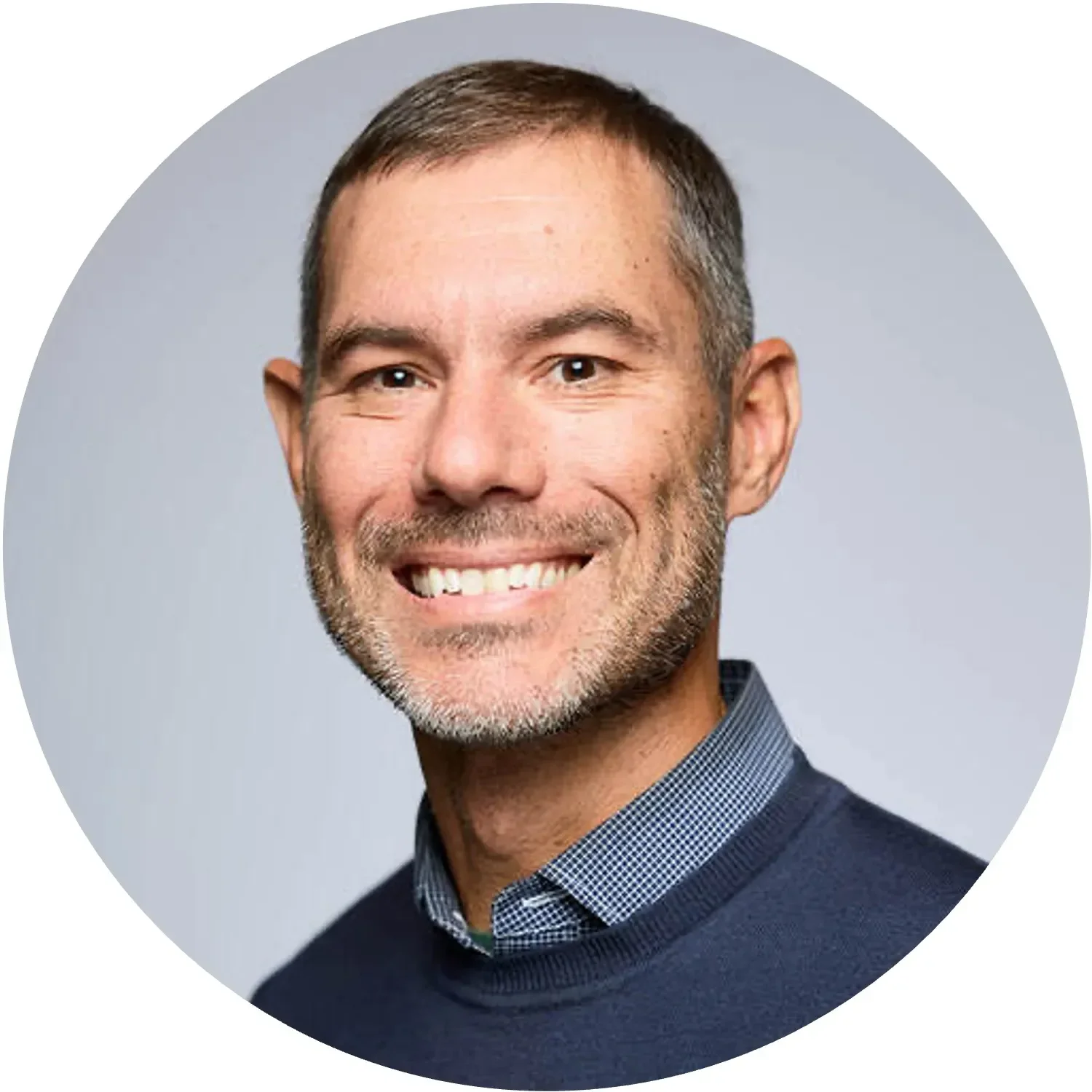 Smiling man with short hair, wearing a navy sweater over a checked shirt, gray background.