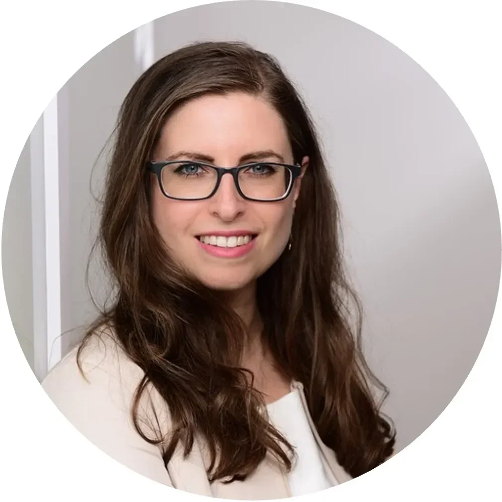 A woman with long brown hair wearing glasses smiles warmly, set against a light background.