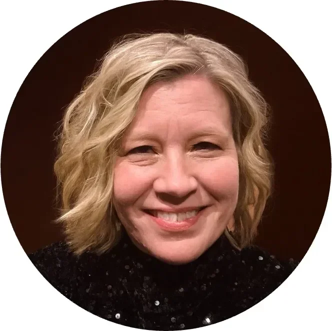 Smiling woman with blonde, curly hair wearing a black top, against a dark background.