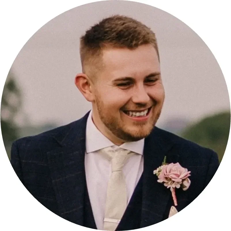 Smiling man in a suit with a pink flower pinned to his lapel, outdoors.