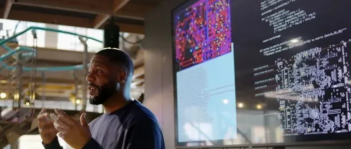 A man gestures while speaking in front of two large digital displays with colorful graphics.