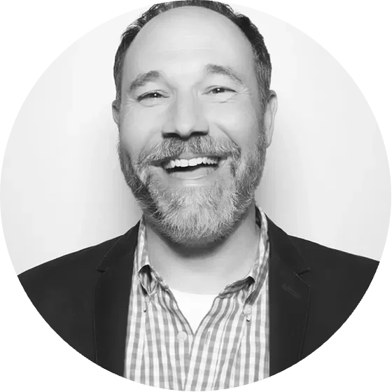 Smiling man with a beard wearing a blazer and plaid shirt against a white background.