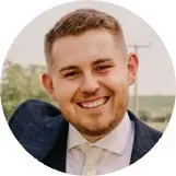 Smiling man in a suit against a blurred outdoor background.