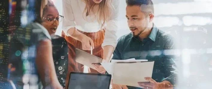 Three professionals collaborate over documents and a laptop in a bright office setting.