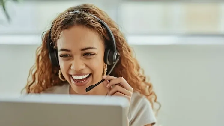 Woman with curly hair smiling while wearing a headset, speaking into a computer.