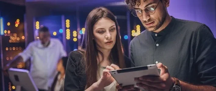 Two people closely examining a tablet, with colorful lights and another person in the background.