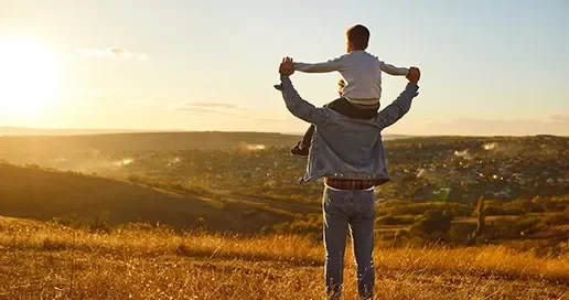 A man holds a child on his shoulders, overlooking a sunset landscape.