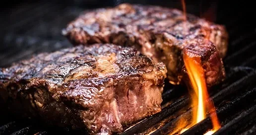 Grilled steaks with charred grill marks and flames in the background.