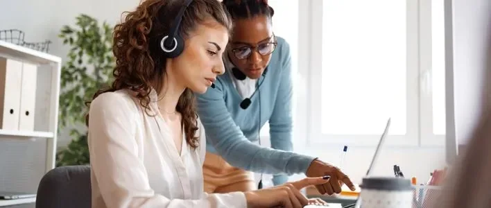 Two women collaborating at a desk; one wears a headset and the other points to a laptop screen.
