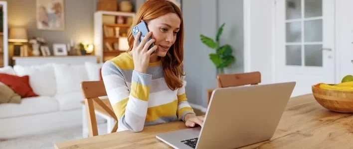 Woman speaking on the phone while using a laptop at a wooden table in a bright room.
