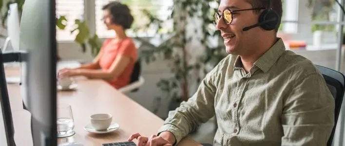 A man with glasses and a headset smiles while working at a computer, a woman sits nearby.