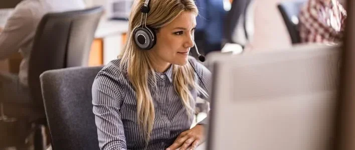 Woman wearing headphones focused on her computer in a busy office setting.