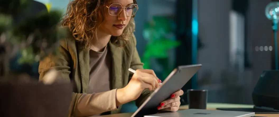 Woman with curly hair wearing glasses, focused on a tablet at a dimly lit desk.