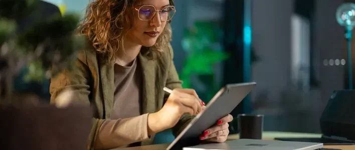 A woman with curly hair and glasses writing on a tablet at a desk with plants in the background.