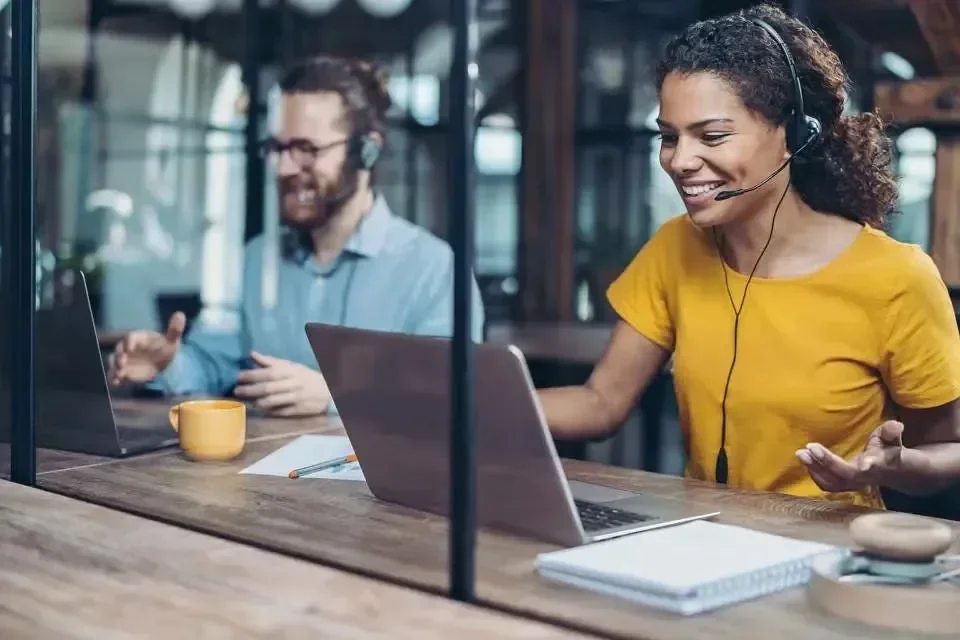 Two people in headsets, smiling and working on laptops in a modern office setting.