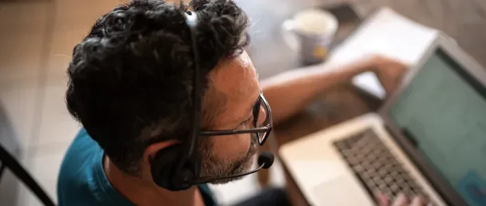 Man with curly hair and glasses wearing a headset, working on a laptop at a table.