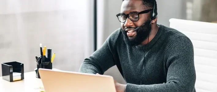 Man wearing a headset, smiling while working on a laptop in a bright office.