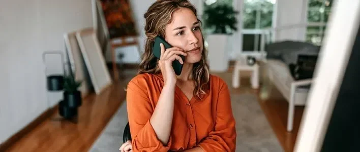 A woman with wavy hair in an orange shirt is talking on a phone in a bright room.