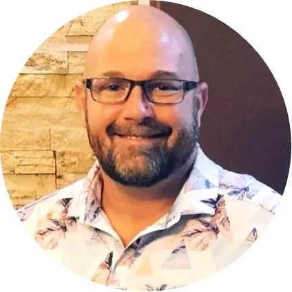 Smiling man with glasses and a floral shirt, against a textured stone background.