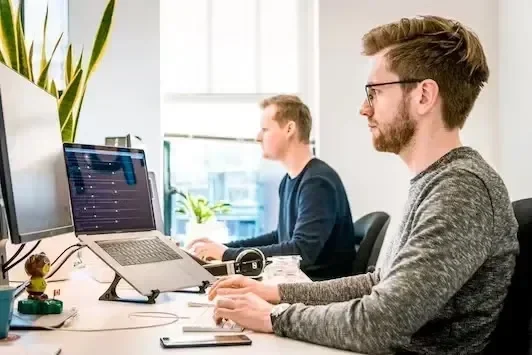 Two men working at desks with multiple computer monitors in a bright office setting.