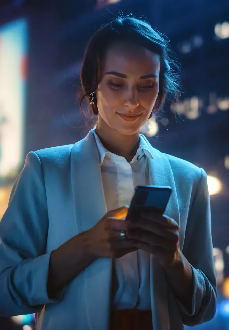 A woman in a light blazer smiles while using her phone at night, illuminated by city lights.