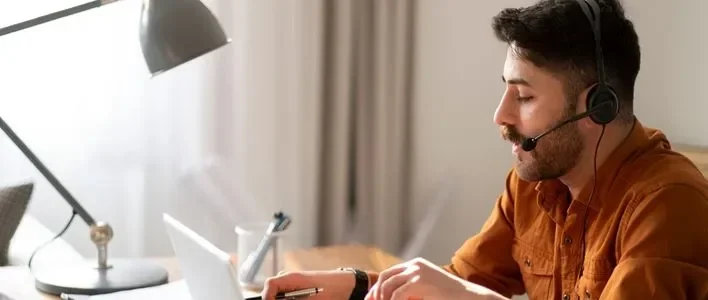 Man with headset wearing orange shirt typing notes on a computer.