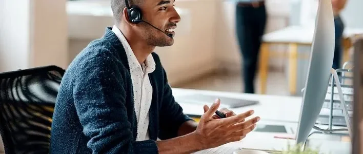 Man in white patterned shirt with blue sweater wearing a headset and gesturing at desktop computer.