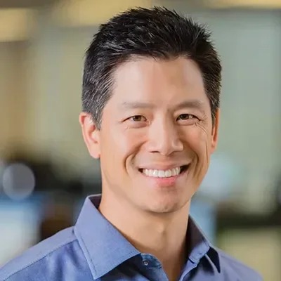 Smiling man in a blue shirt with short dark hair and a blurred office background.