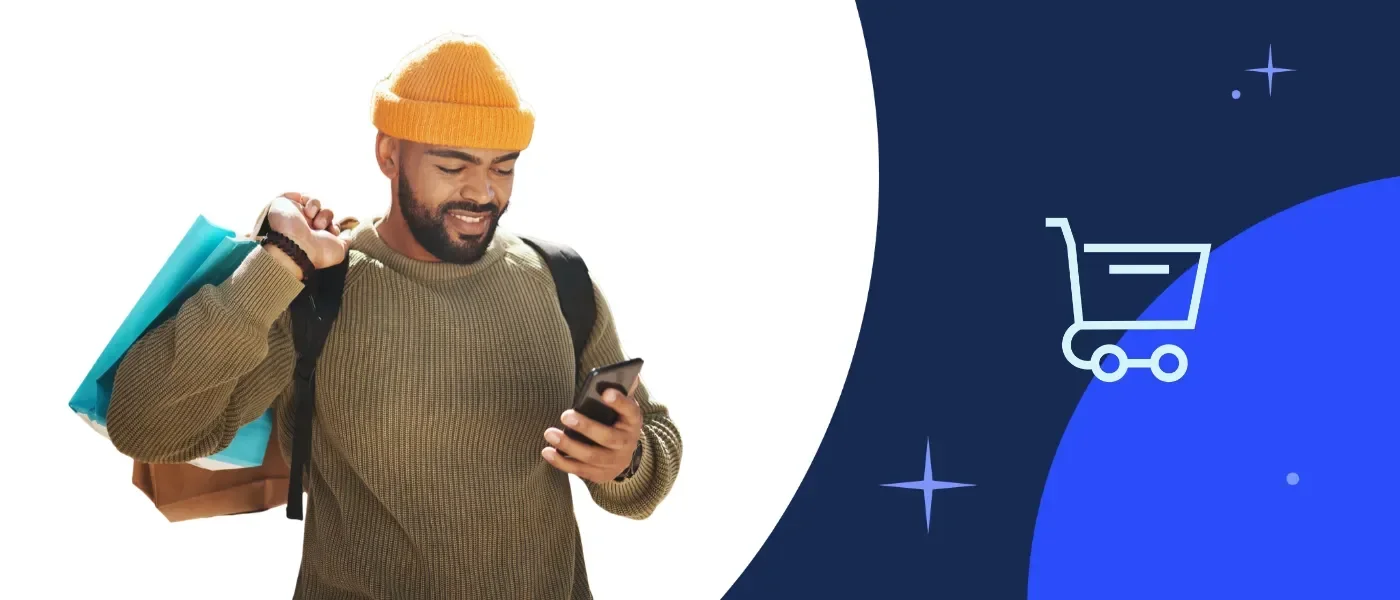 Man in a sweater and yellow beanie smiles while checking his phone, carrying shopping bags.