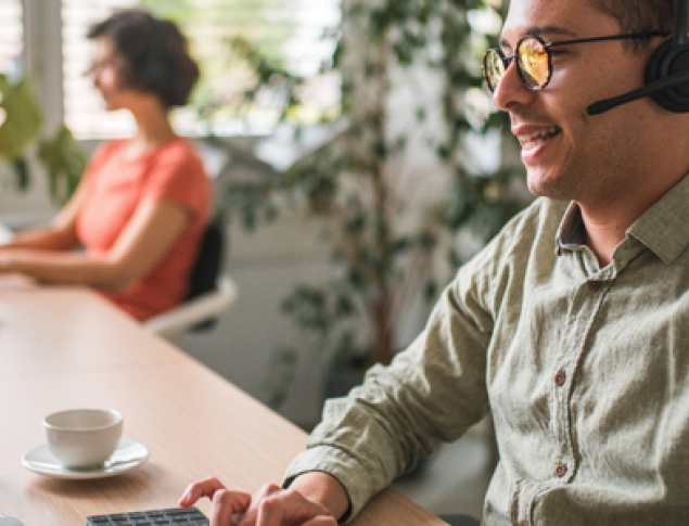 A smiling man wearing headphones in a contact center with a woman in the background.